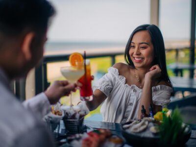A woman in a white off-the-shoulder top smiles while toasting with cocktails at a restaurant table with seafood and snacks by the ocean at Tides Coastal Kitchen.