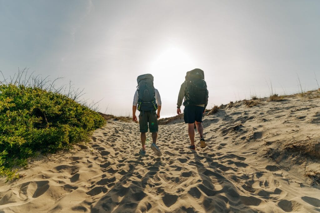 Two hikers with backpacks walk along a sandy path towards the sun, surrounded by coastal vegetation in False Cape State Park.