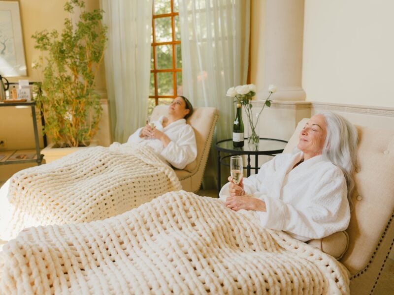 Two women relax in plush chairs at spa, wrapped in chunky knit blankets, each holding a glass of sparkling drink at Flowering Almond Spa in The Founders Inn.