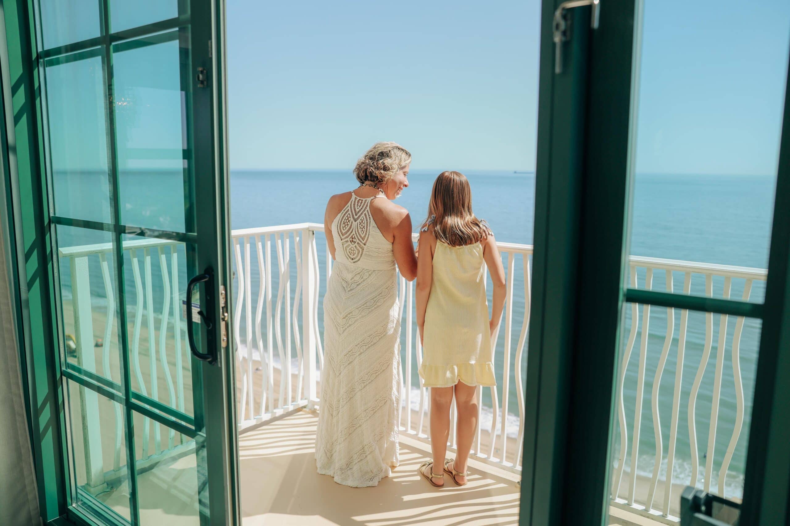 A mom and a daughter stand together on a balcony overlooking the ocean, enjoying a sunny day with clear blue skies at the Hilton Oceanfront.