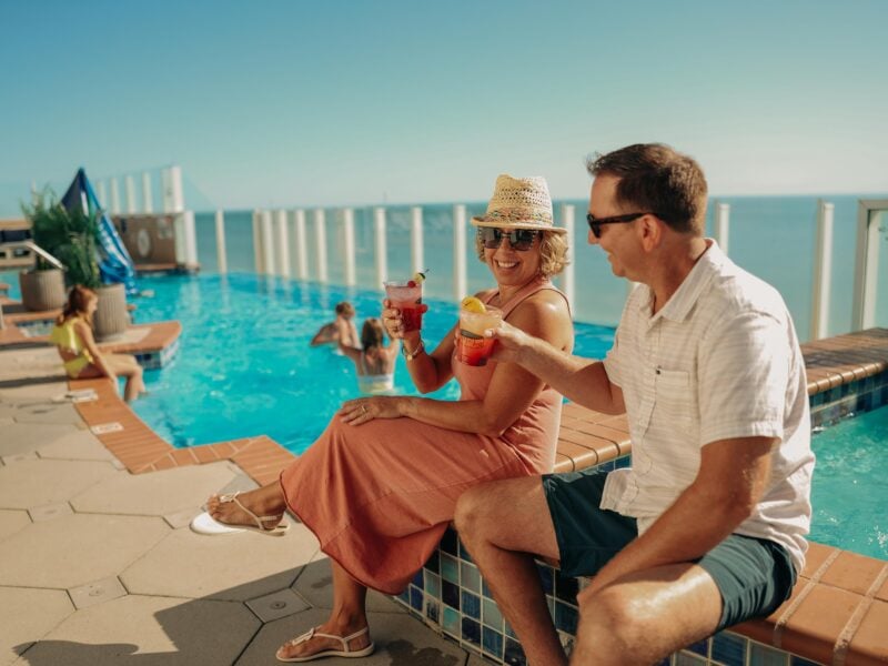 Couple enjoying colorful cocktails by a roof top pool under a clear blue sky, kids playing in a pool, and vibrant scenery at Hilton Oceanfront.