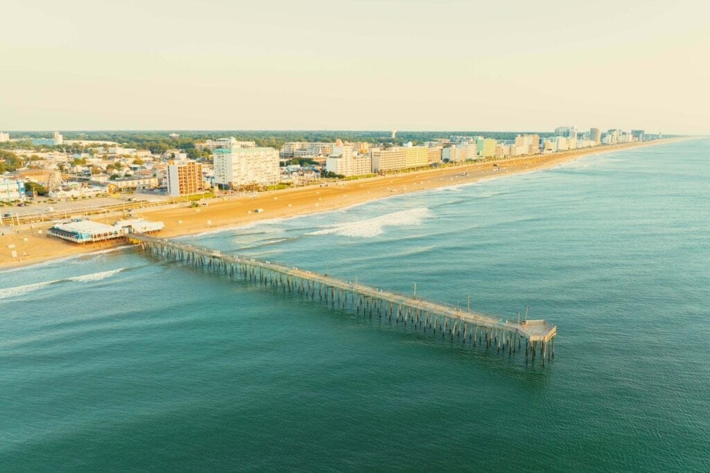 Aerial view of Virginia Beach Fishing Pier extending into calm waters, with sandy beach and beachfront hotels in the background at the Oceanfront.