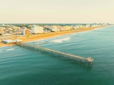 Aerial view of Virginia Beach Fishing Pier extending into calm waters, with sandy beach and beachfront hotels in the background at the Oceanfront.