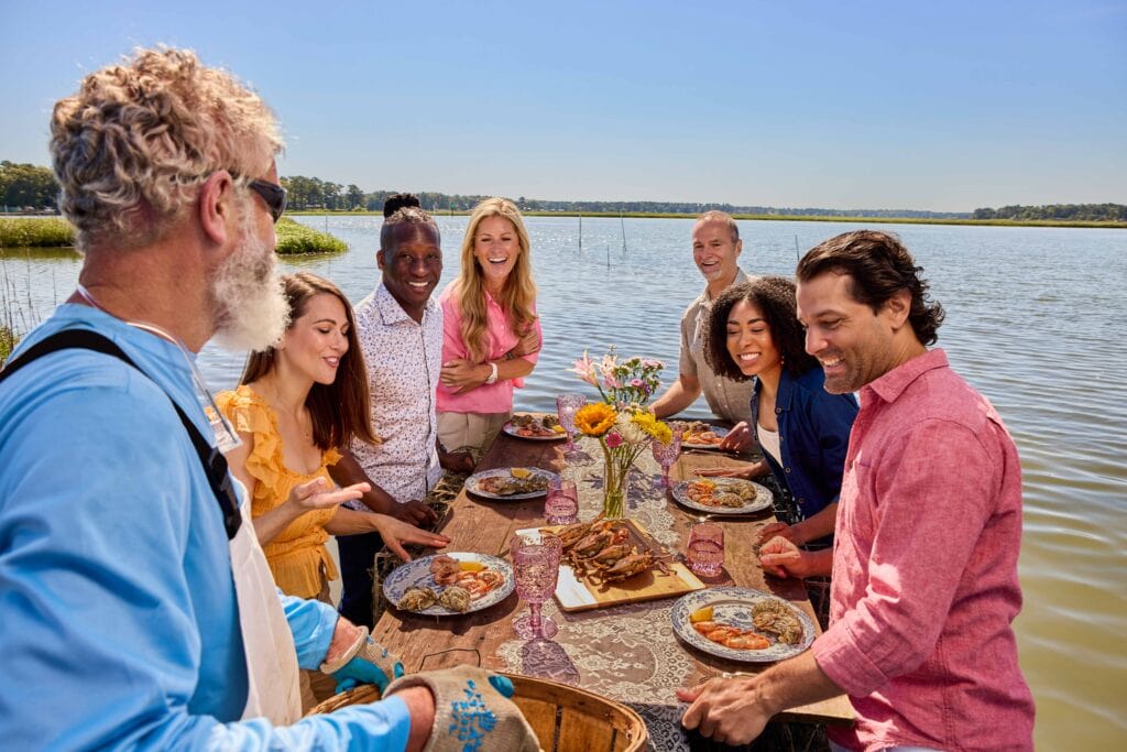A group of friends enjoying a seafood feast outdoors the scenic waterfront of the Chesapeake Bay, surrounded by vibrant flowers and sunlight in the Spring.