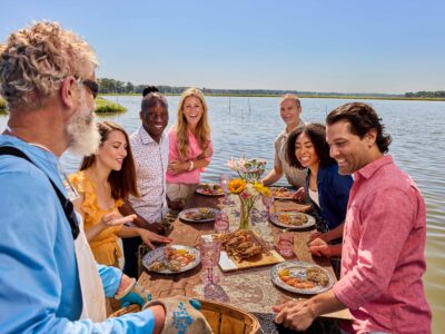 A group of friends enjoying a seafood feast outdoors the scenic waterfront of the Chesapeake Bay, surrounded by vibrant flowers and sunlight in the Spring.