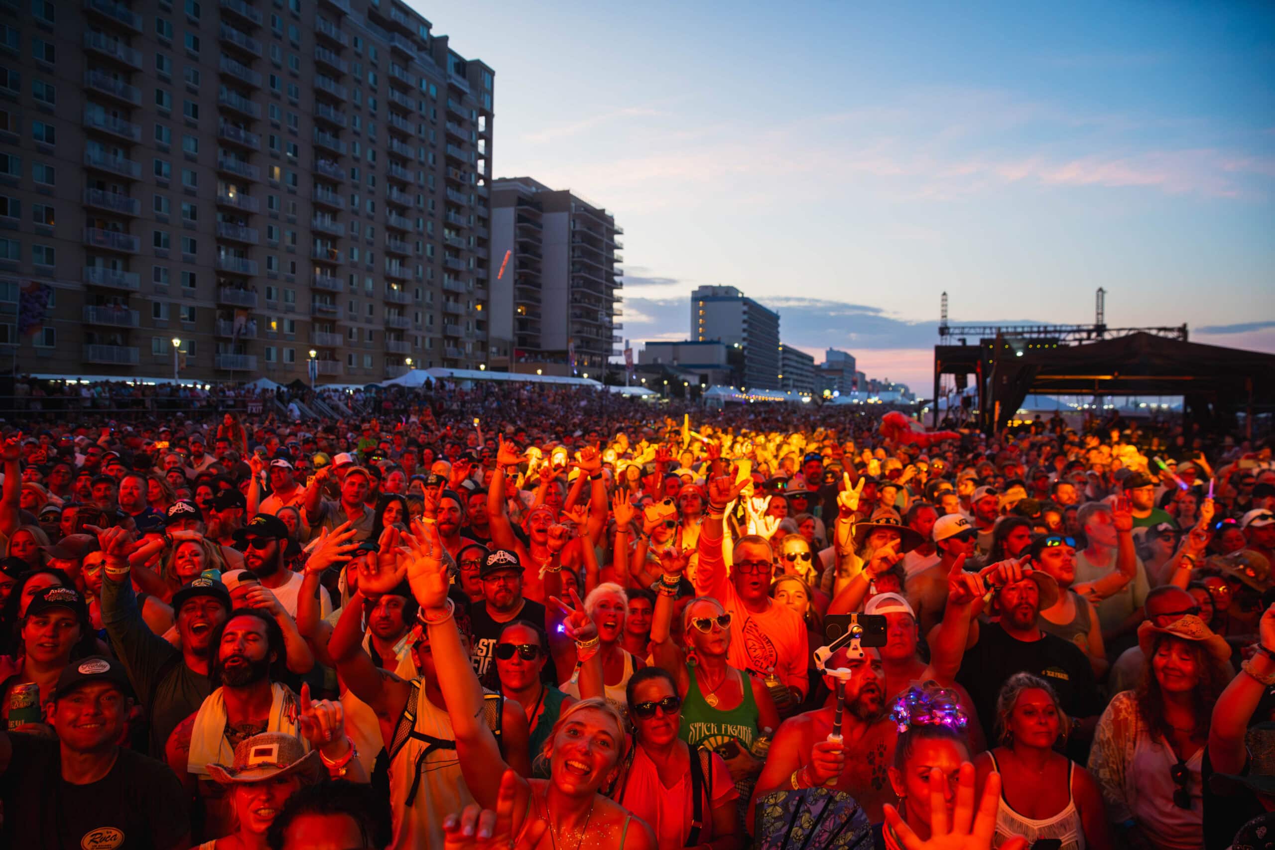 A vibrant crowd at Point Music Festival on the Virginia Beach Oceanfront, with people cheering and waving under a colorful sunset and city skyline backdrop.