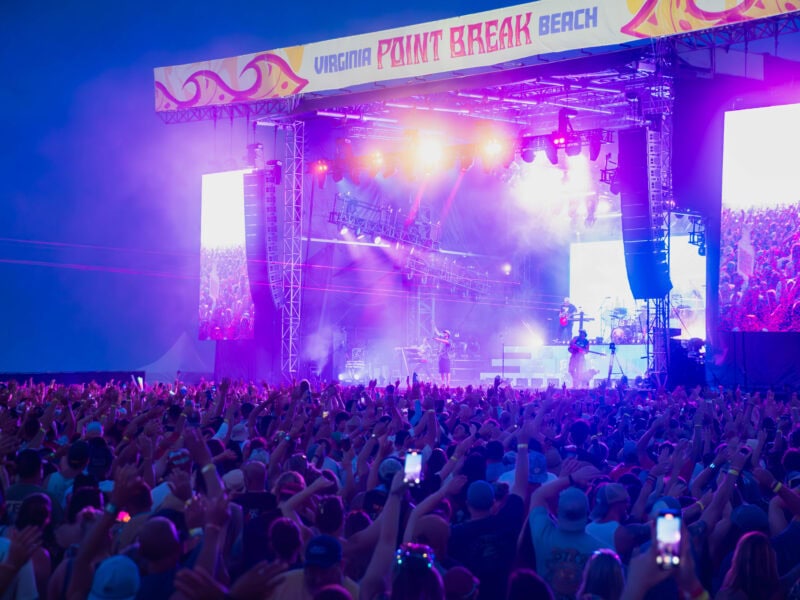 A vibrant concert scene at Point Break Music Festival in Virginia Beach, featuring a large crowd with raised hands and colorful stage lights.