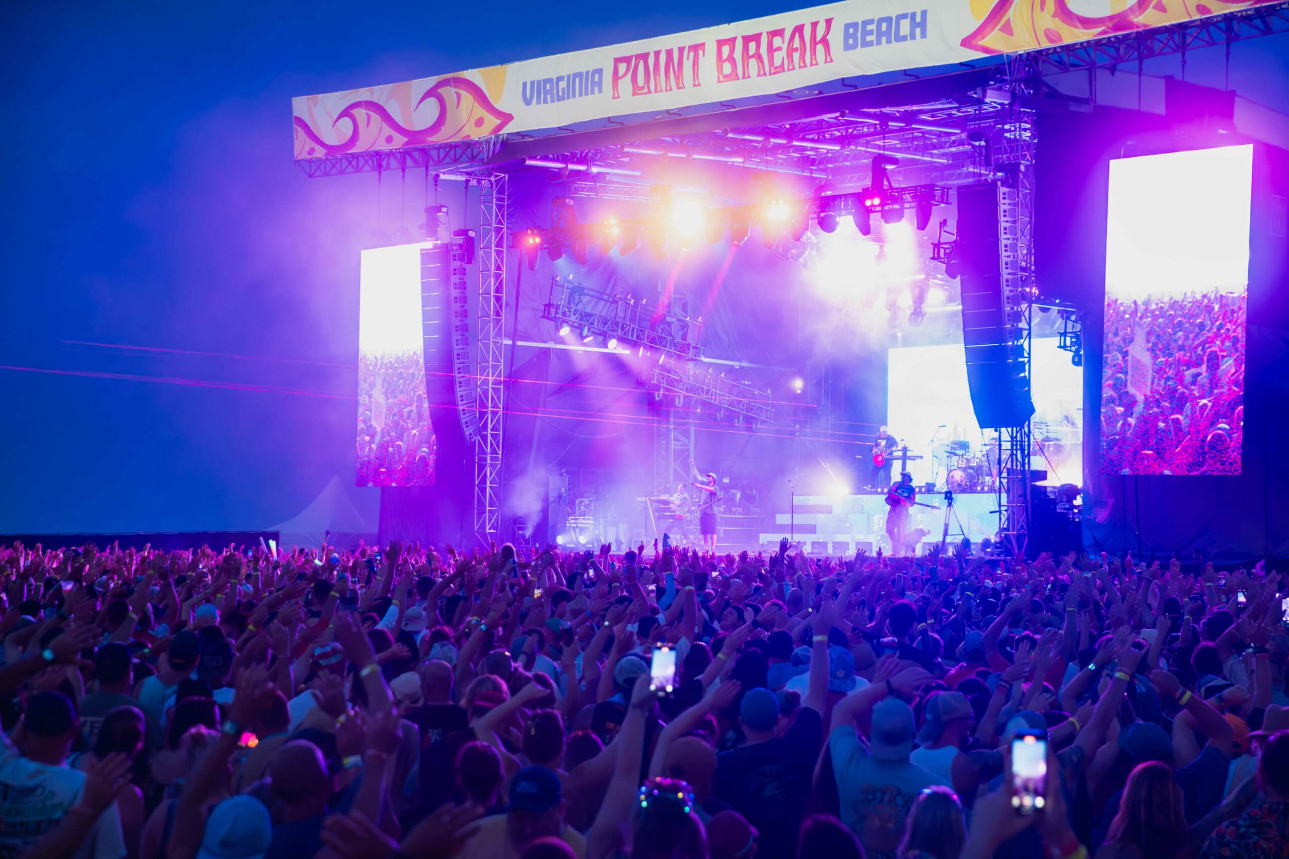 A vibrant concert scene at Point Break Music Festival in Virginia Beach, featuring a large crowd with raised hands and colorful stage lights.