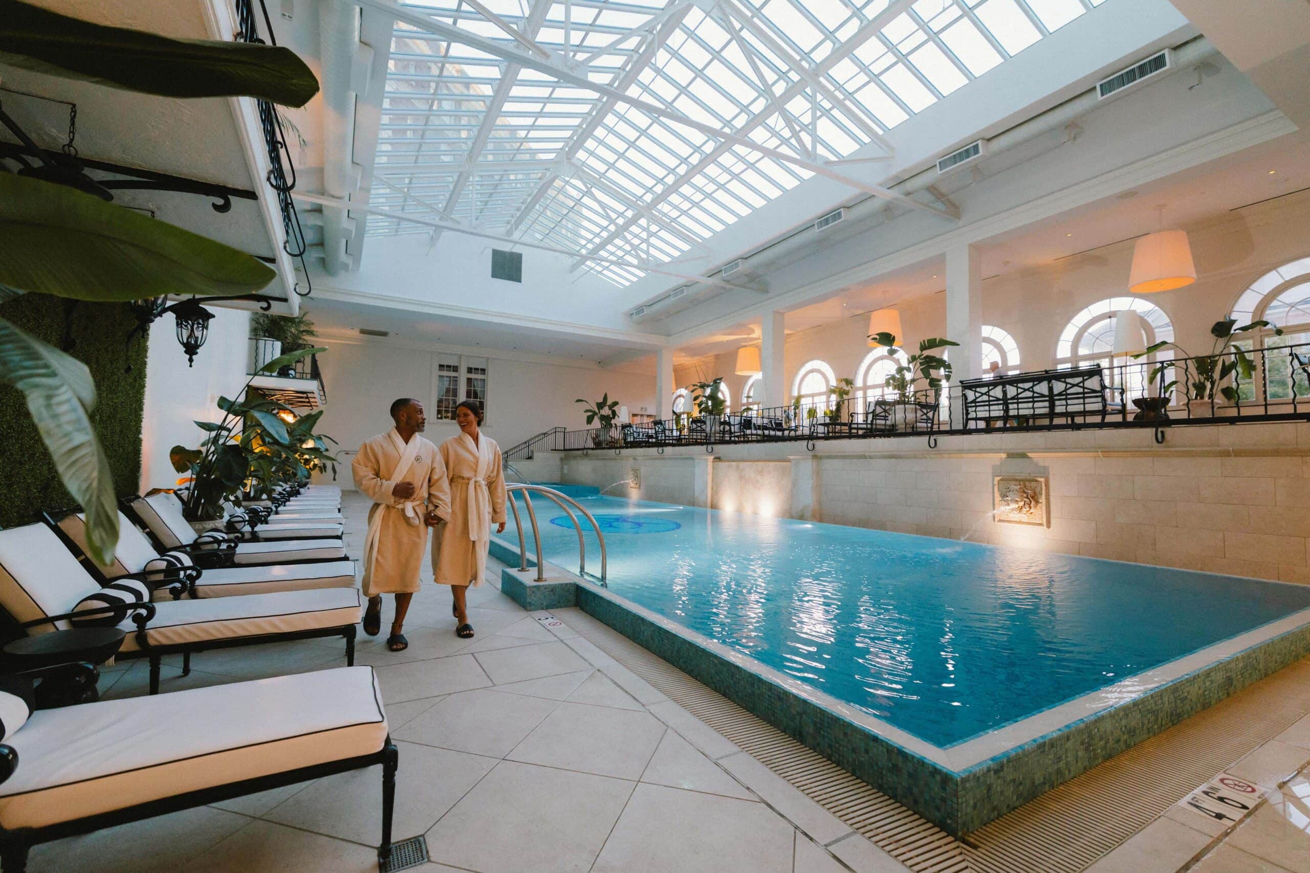A couple in bathrobes walk hand-in-hand by a luxurious indoor pool surrounded by plants and elegant decor at The Historic Cavalier.