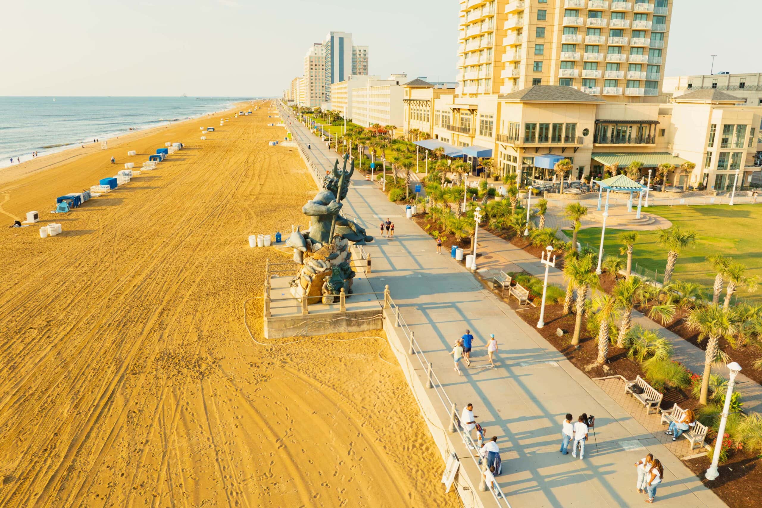 Aerial view of the Virginia Beach boardwalk with people walking, a statue, palm trees, and beachfront hotels at sunset.