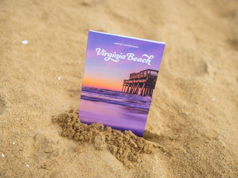 A Virginia Beach official visitors guide partially buried in sand, featuring a colorful cover with a pier at sunset in.