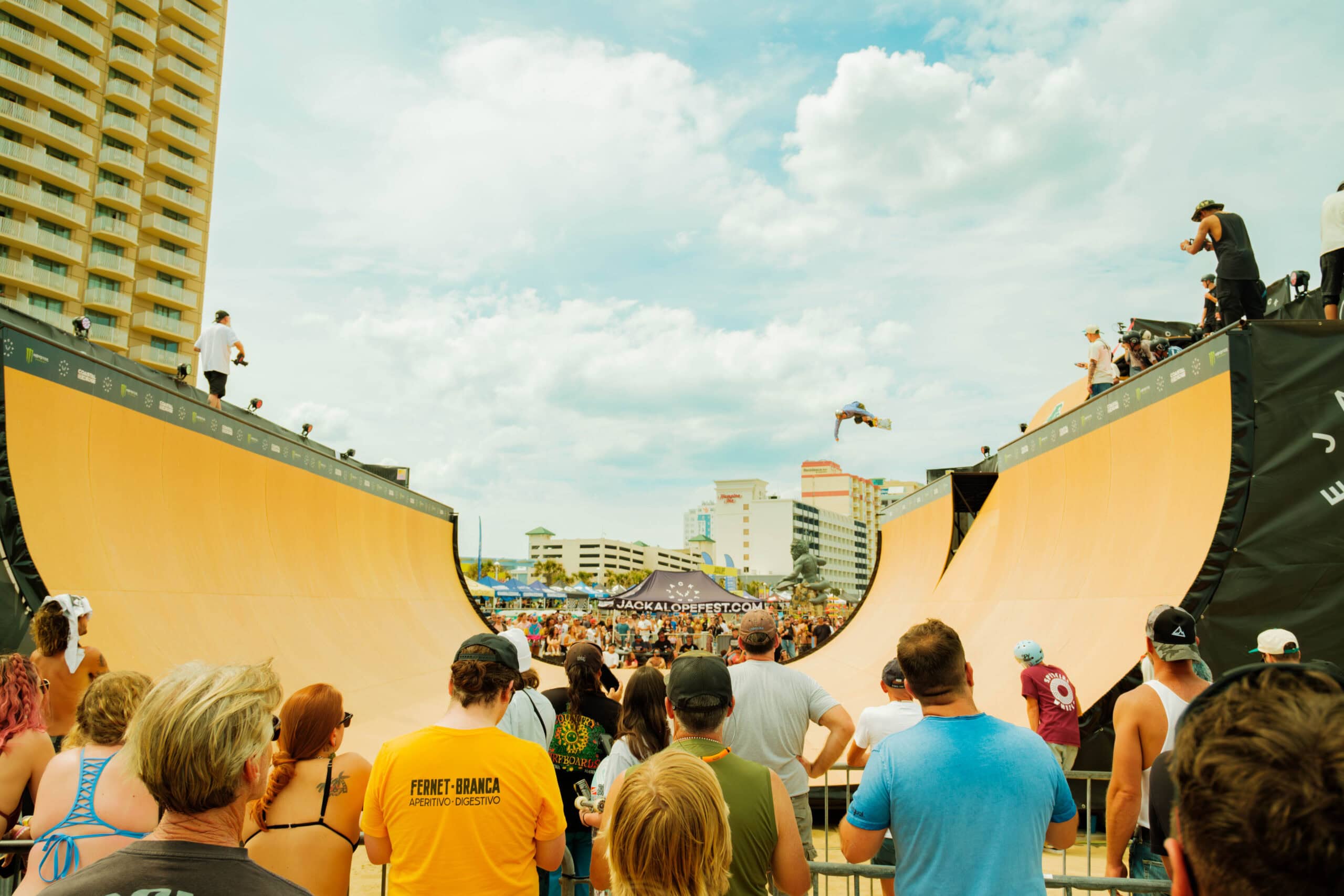 A skateboarder performs a trick in mid-air above a large ramp during the lively Jackalope Festival, surrounded by an enthusiastic crowd.