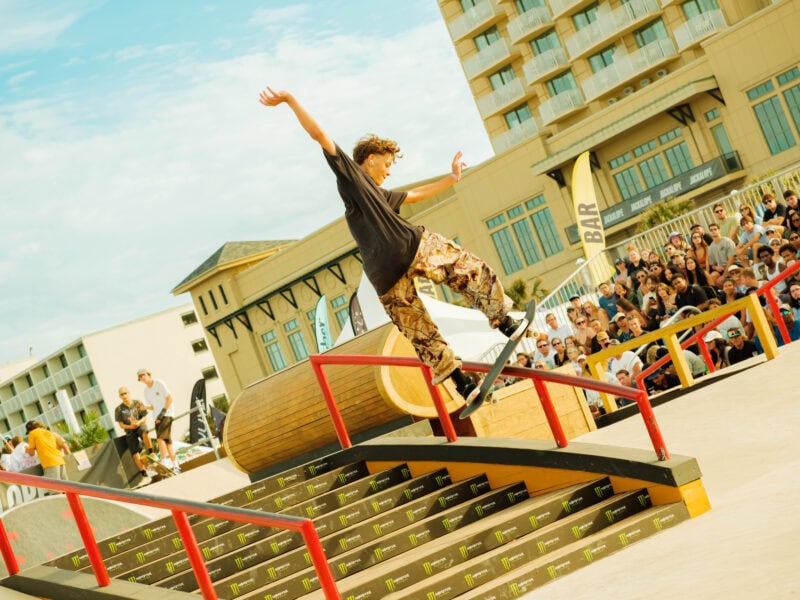 A skateboarder performs a trick on a rail at a crowded skateboarding event, with spectators watching in the background.