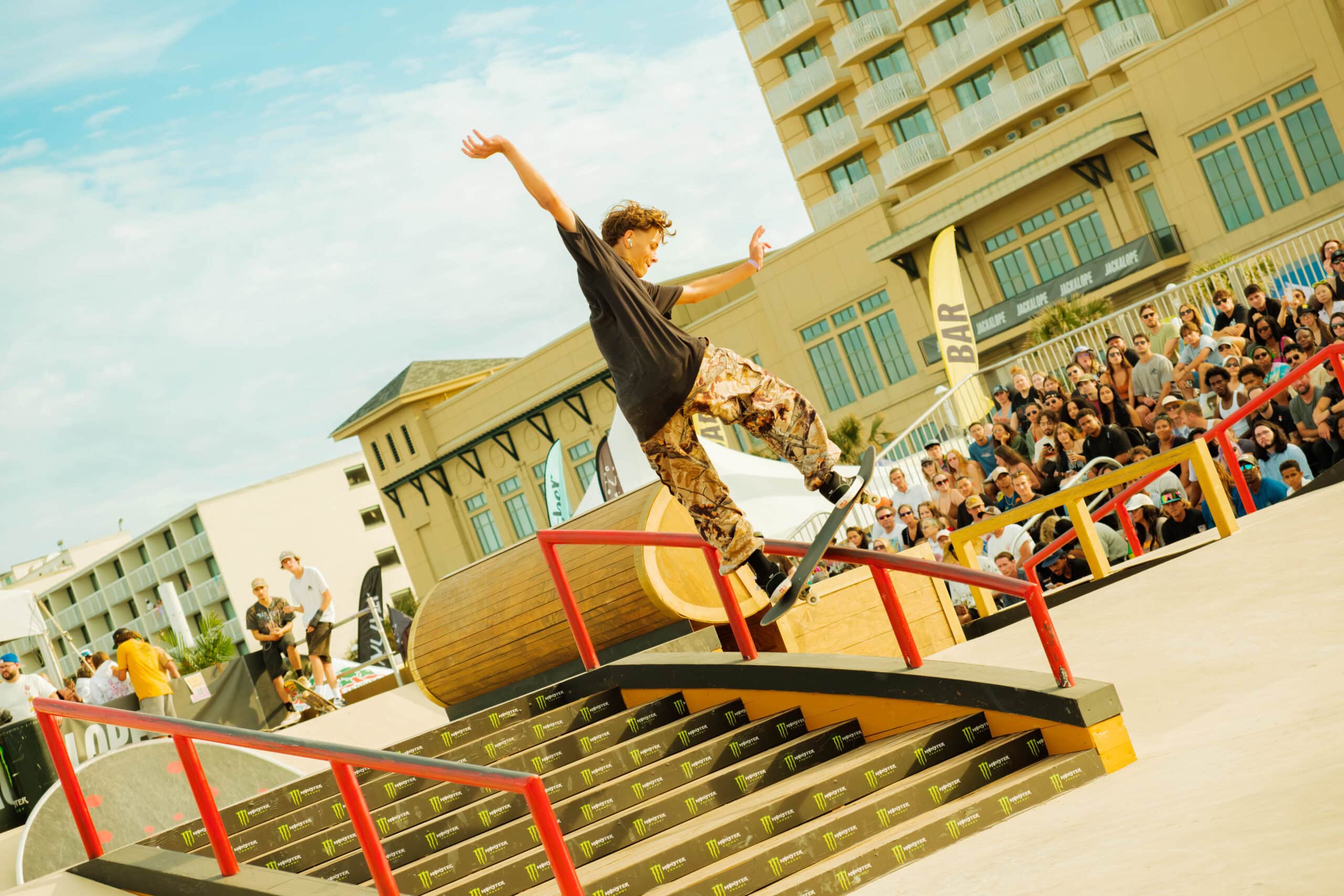 A skateboarder performs a trick on a rail at a crowded skateboarding event, with spectators watching in the background.