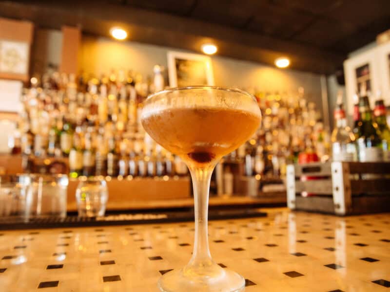 A close-up of a cocktail in a glass with a cherry, set against a blurred bar filled with bottles.