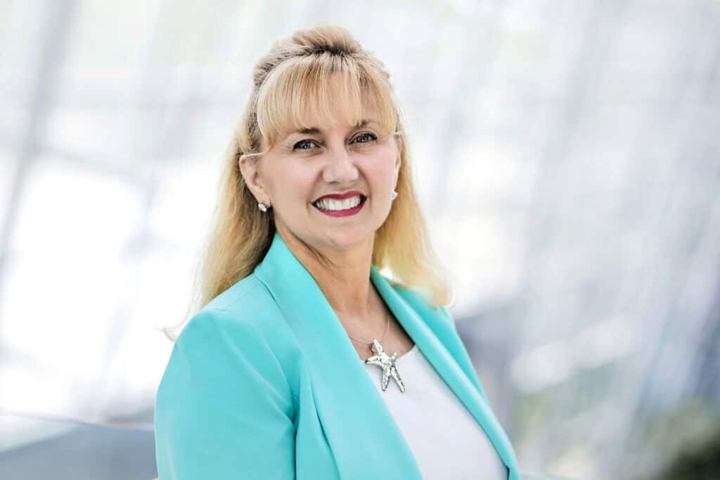 A smiling woman with blonde hair, wearing a turquoise blazer and silver jewelry, poses against a softly blurred background.