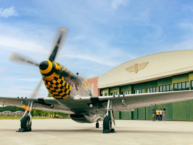 A vintage aircraft with a checkered nose is parked outside a hangar at the Military Aviation Museum while a group of people stands nearby, looking at it.