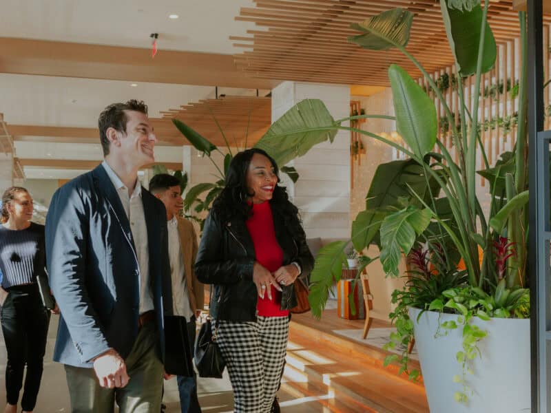 A diverse group of four individuals, dressed in casual business attire, smiles and engages while walking through a modern, plant-filled interior.
