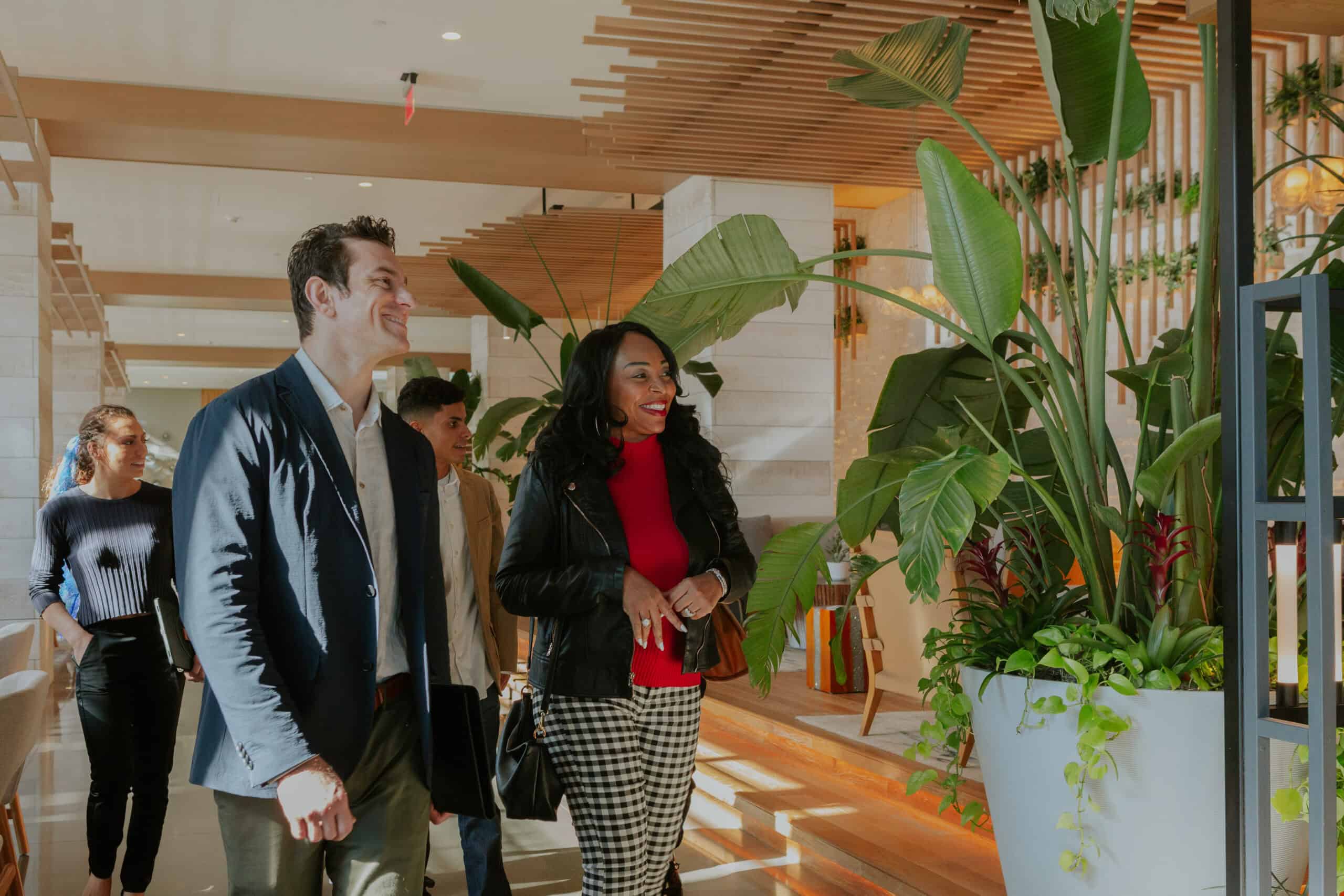 A diverse group of four individuals, dressed in casual business attire, smiles and engages while walking through a modern, plant-filled interior.