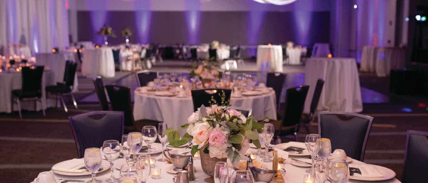 Elegant banquet setup featuring round tables adorned with floral arrangements, candles, and glassware, set in a softly lit venue.