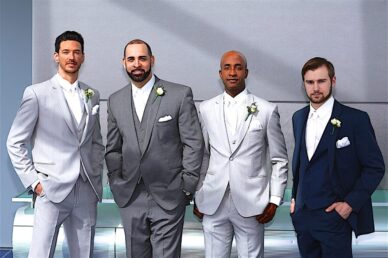 Four well-dressed men in formal suits, two in light gray and two in navy, each wearing a boutonnière, stand in a modern setting.