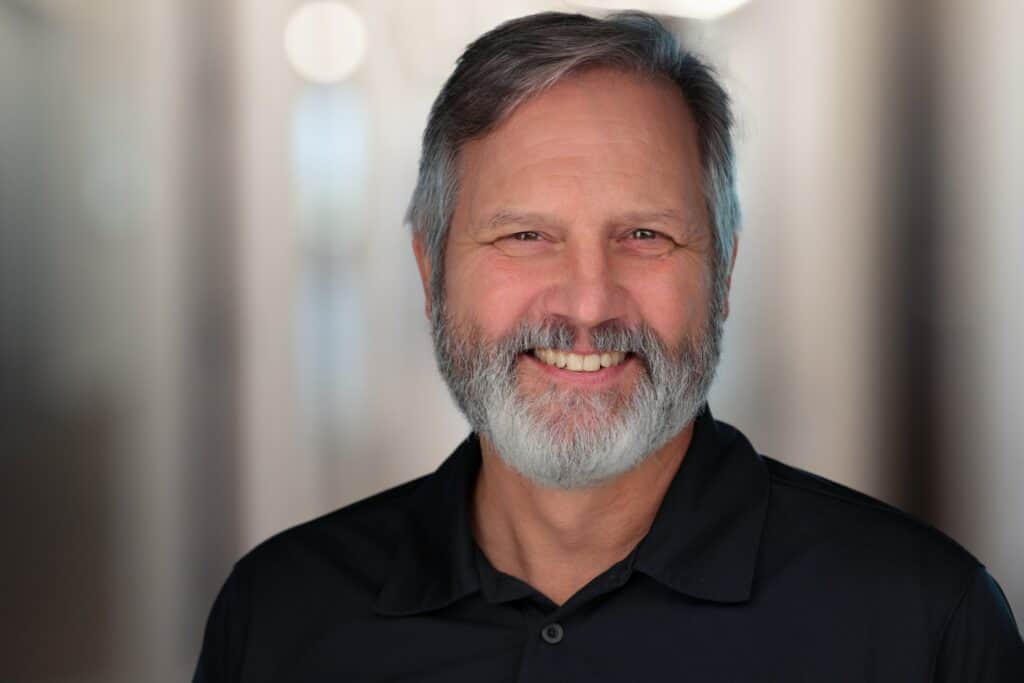 Smiling middle-aged man with a beard wearing a black polo shirt, set against a softly blurred background.