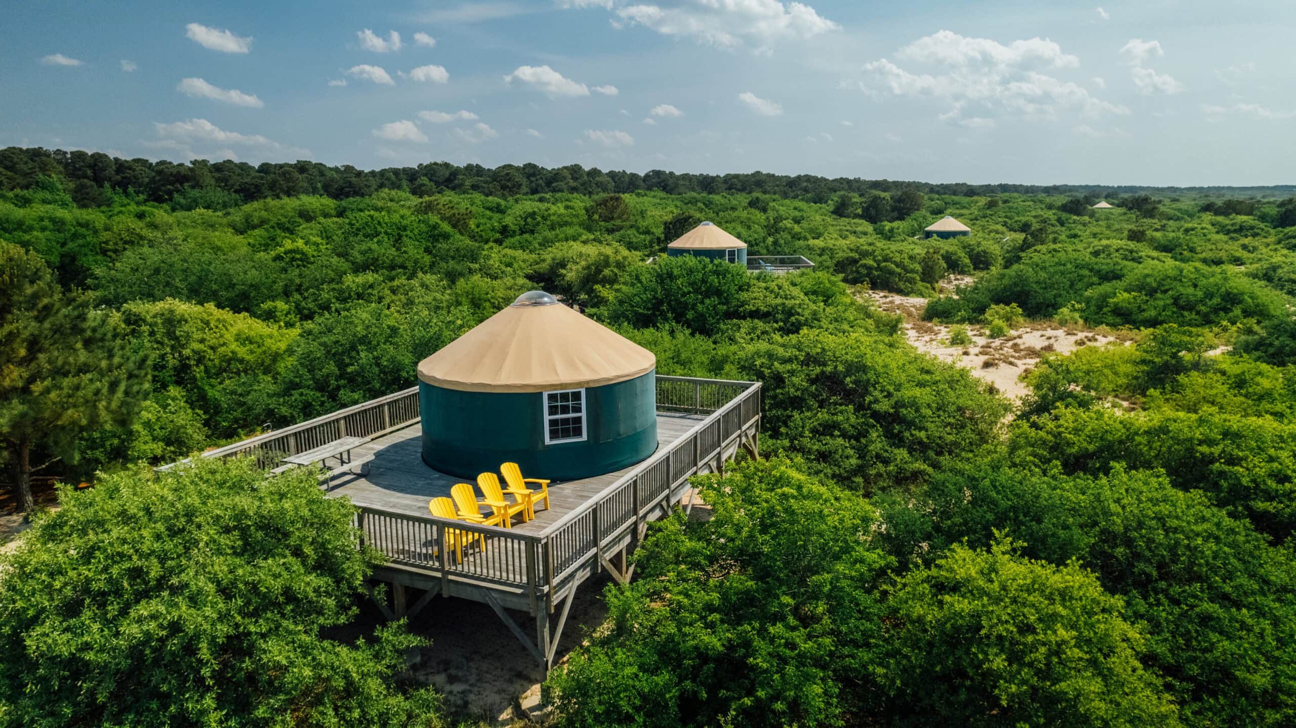 Aerial view of round, green yurts with tan roofs on a raised deck, surrounded by lush greenery and trees under a blue sky at First Landing State Park.