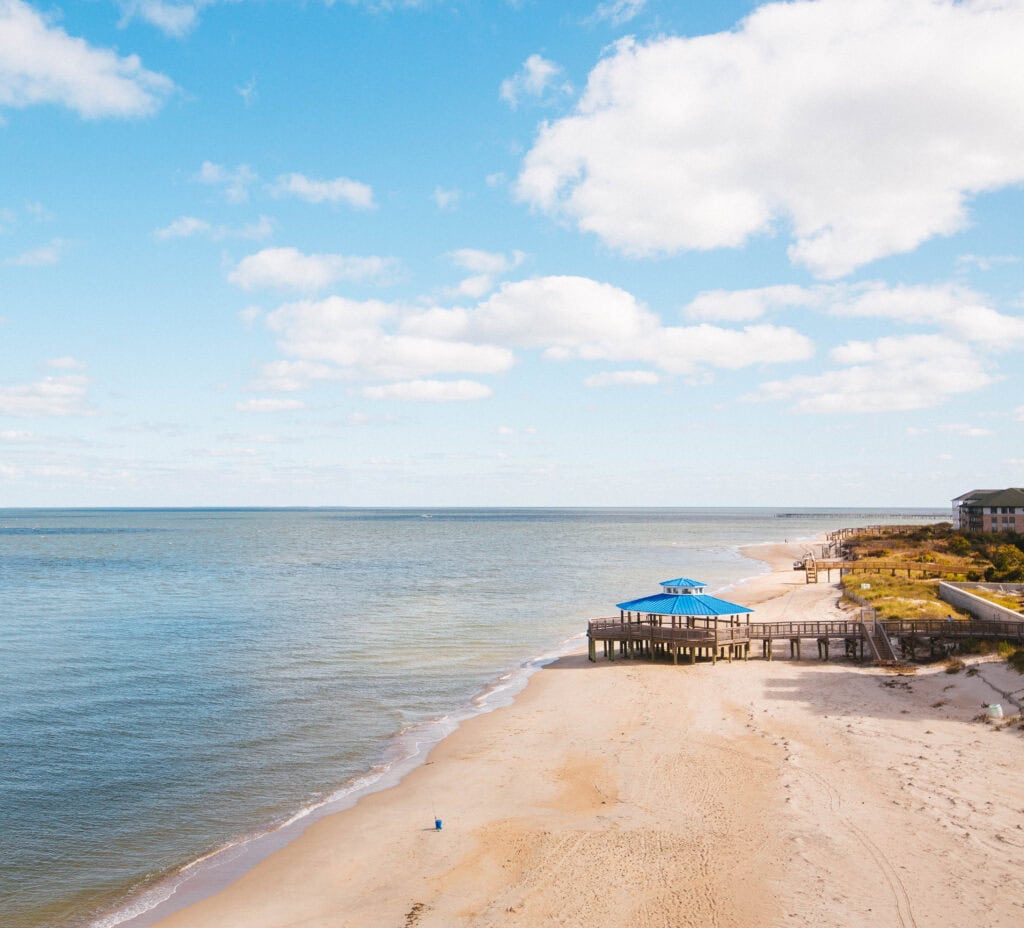 A tranquil beach scene featuring a wooden pier with a blue roof, soft golden sand, and a clear, sunny sky with fluffy clouds.