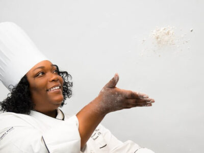 A smiling female chef in a white uniform and tall hat playfully tosses flour into the air against a light background.