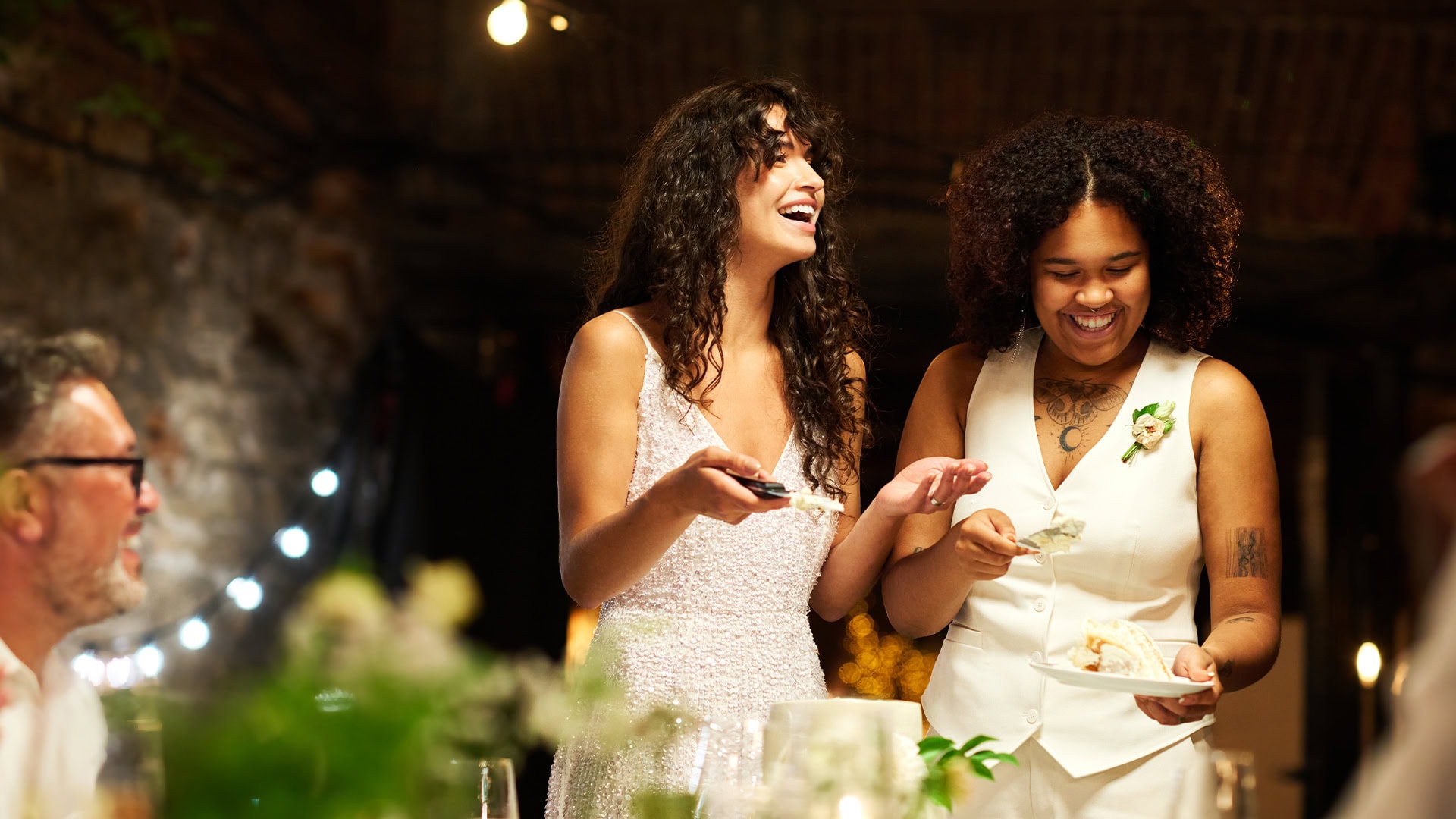 Two brides are enjoying slicing into their wedding cake surrounded by smiling family.