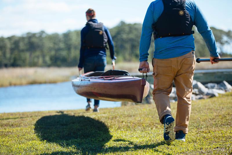 A couple both dressed in long sleeves and pants with backpacks on are carrying a kayak toward a lake in False Cape State Park while carrying oars in their other hand.