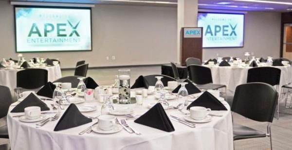 A banquet hall is pictured containing tables with white table cloths, place settings and black triangular cloth napkins and in the background two TVs are mounted to the wall displaying the words "Apex Entertainment."