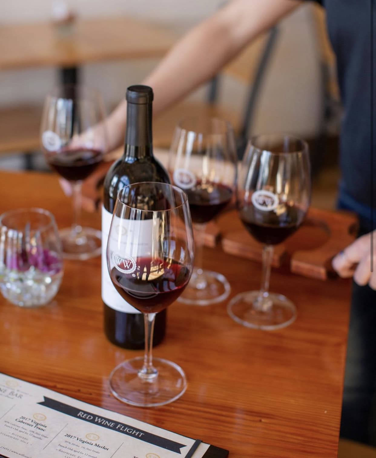At Williamsburg Winery four wine glasses containing a red wine surround a bottle of wine set out on a table in front of a menu. A person can be seen blurred in the background with their hands leaning on the table.