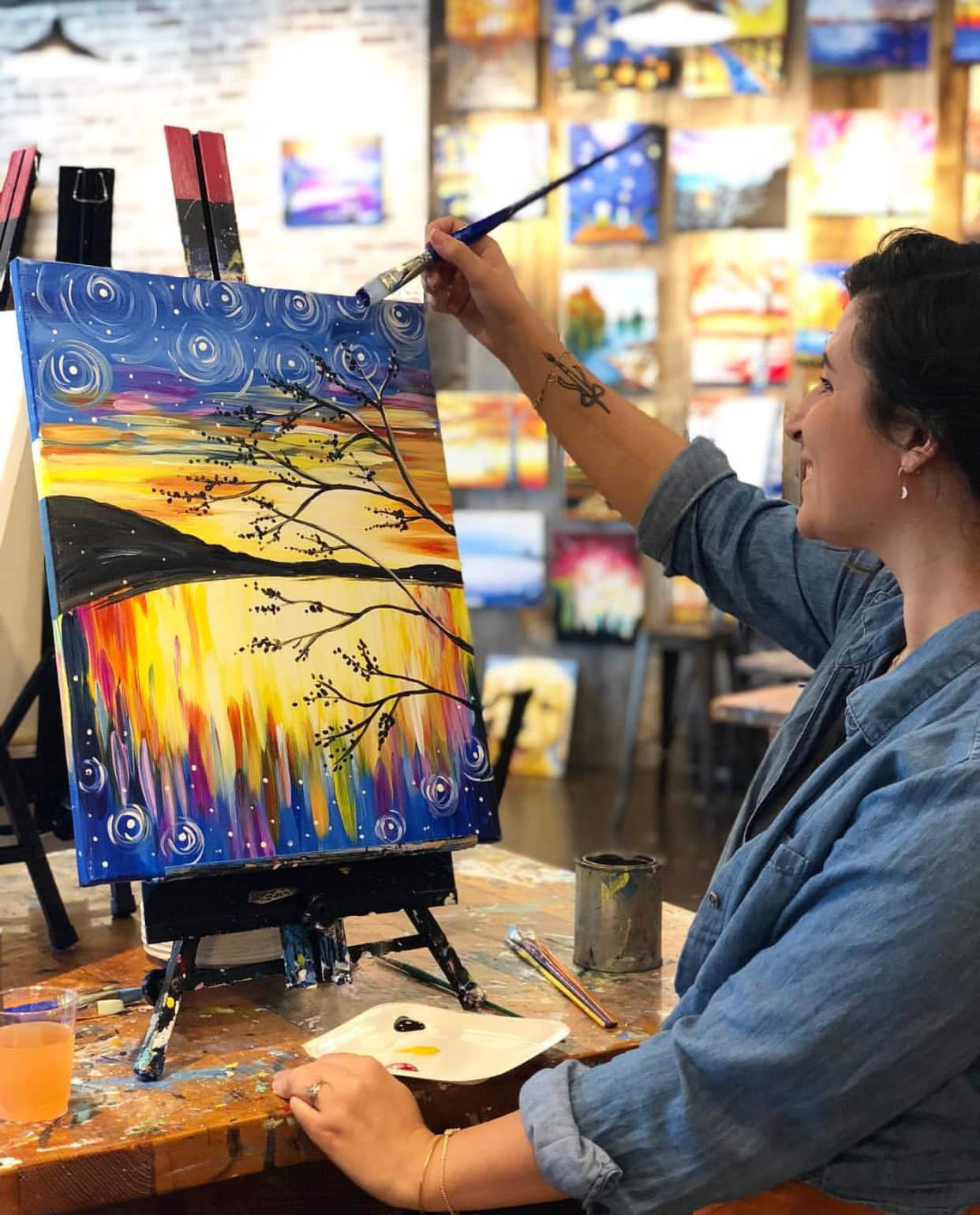 At Muse Paint Bar, a smiling woman in a denim shirt cuffed at the sleeves is painting a colorful landscape on a canvas.