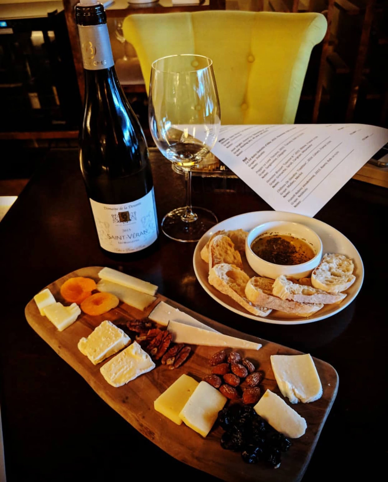 At Tinto Wine & Cheese there is a table with a platter of cheese, nuts, and fruit, a bowl of dip with pieces of bread and a bottle of wine with a wine glass next to it. There is a bright yellow chair pulled up to the table with a white menu in the background.