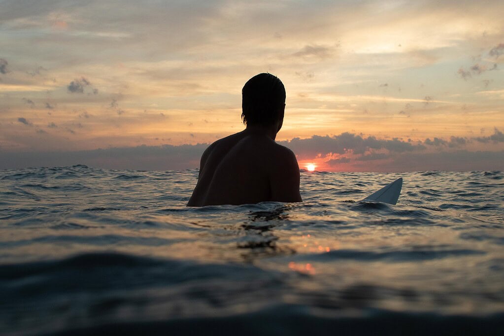 A man sits on a surfboard in chest high waters in the ocean with a vibrant sunrise in the background in Virginia Beach.