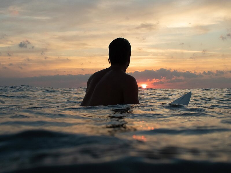 A man sits on a surfboard in chest high waters in the ocean with a vibrant sunrise in the background in Virginia Beach.