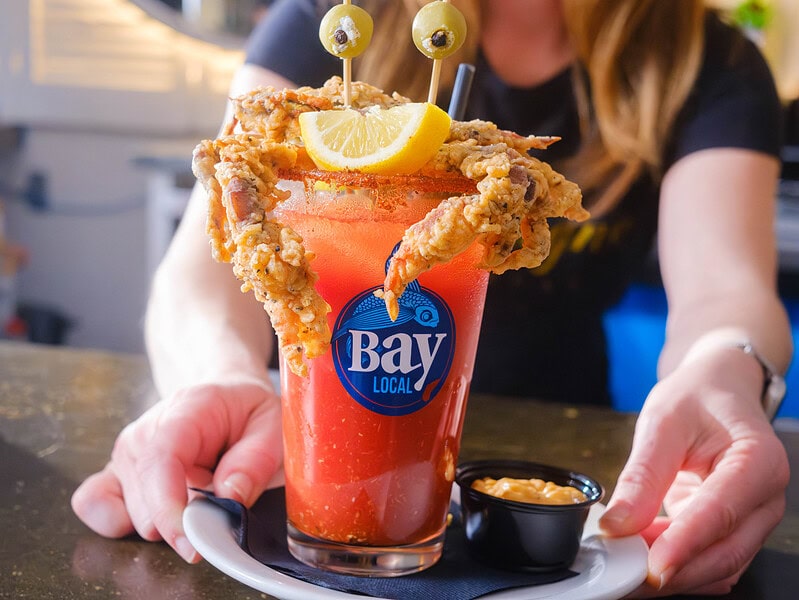 A woman serving a vibrant colored Bloody Mary on a plate with fried crab legs, olives, and lemon garnish on top.
