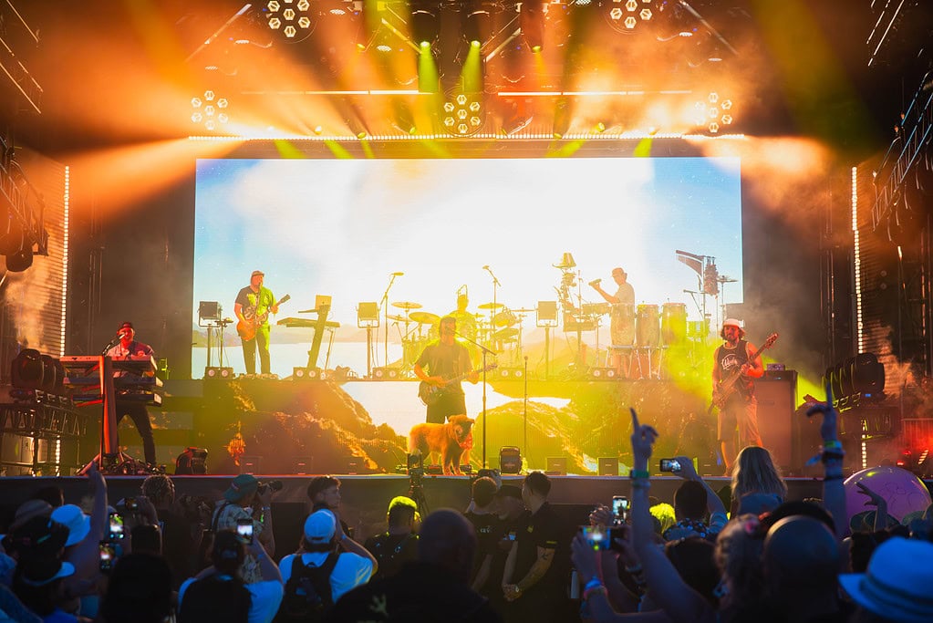 An energetic band on stage with a large screen behind them with vibrant orange and yellow lighting casting on the band and excited crowd at Point Break Music Festival.