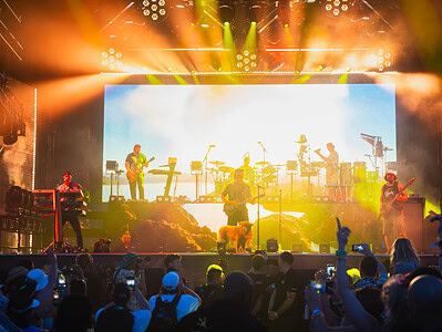 An energetic band on stage with a large screen behind them with vibrant orange and yellow lighting casting on the band and excited crowd at Point Break Music Festival.