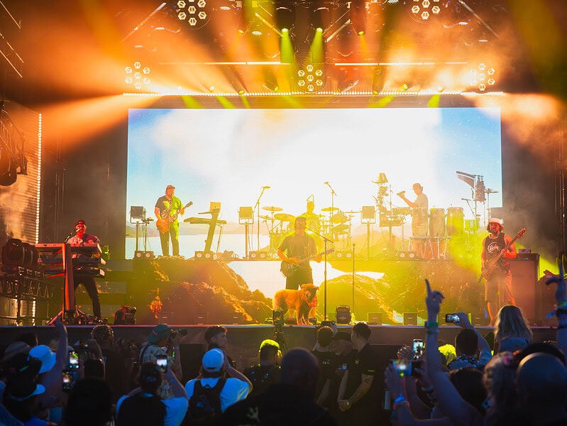 An energetic band on stage with a large screen behind them with vibrant orange and yellow lighting casting on the band and excited crowd at Point Break Music Festival.