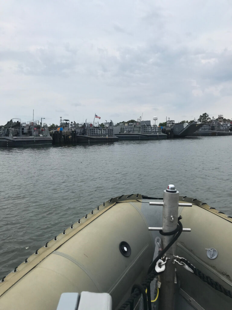 A view from a boat showing a harbor with several military ships docked under a cloudy sky.