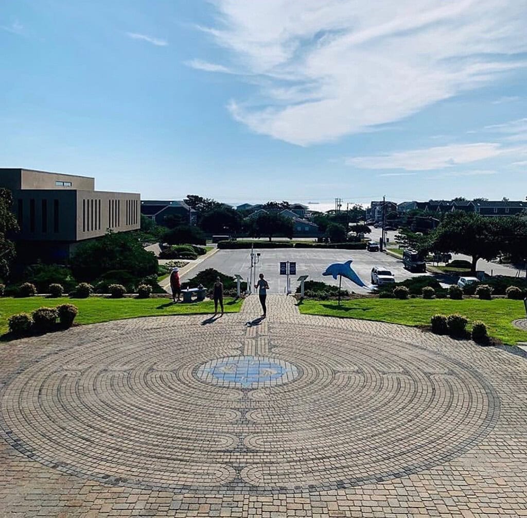Wide view of the circular stone labyrinth at the Edgar Cayce A.R.E. campus with a pattern, surrounded by a grassy area. Three people walk in the distance under a partly cloudy sky. Buildings and a parking lot are visible.