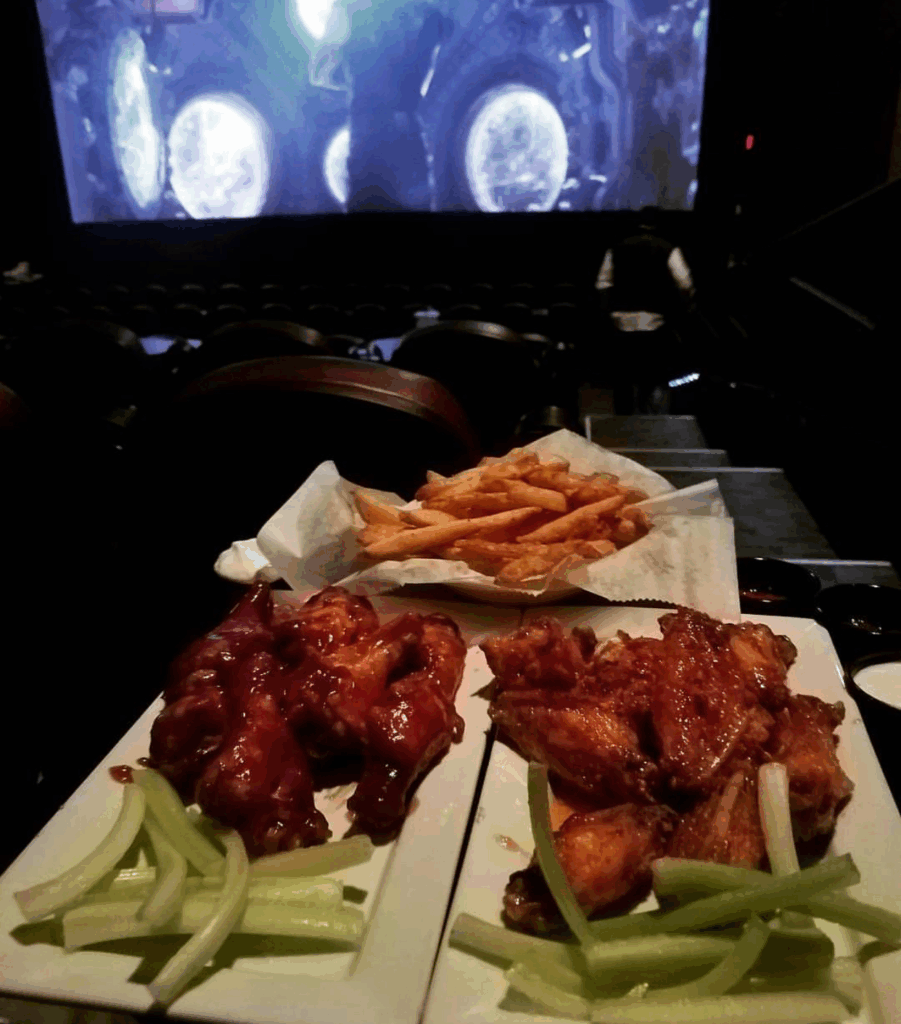Two plates of chicken wings and celery sticks with a basket of fries in a dimly lit movie theater.
