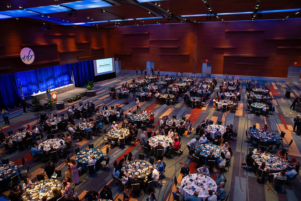 A full ballroom with rich wood panel walls & vibrant blue ceiling lighting with decorated tables and stage with attendees and a speaker