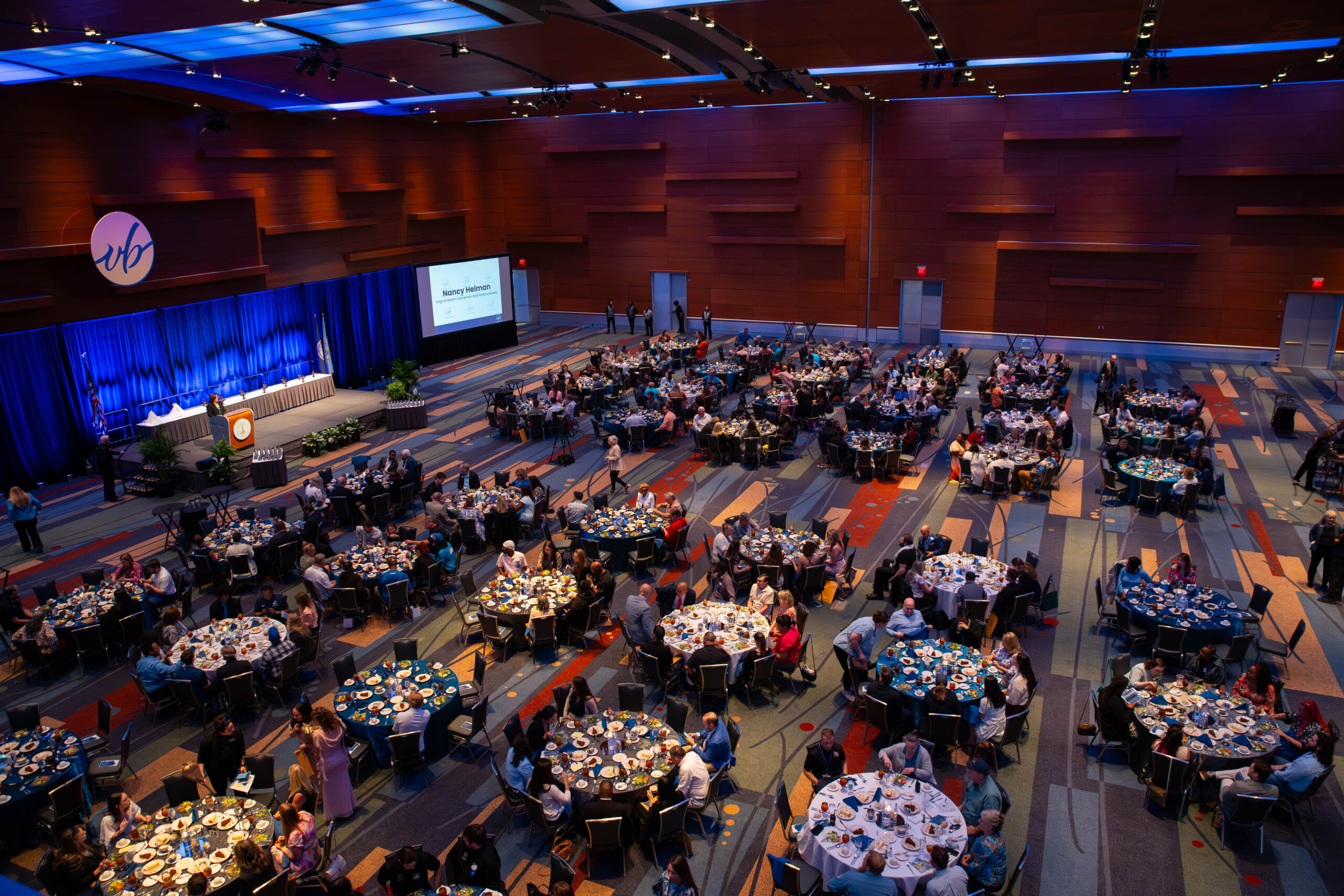 A full ballroom with rich wood panel walls & vibrant blue ceiling lighting with decorated tables and stage with attendees and a speaker