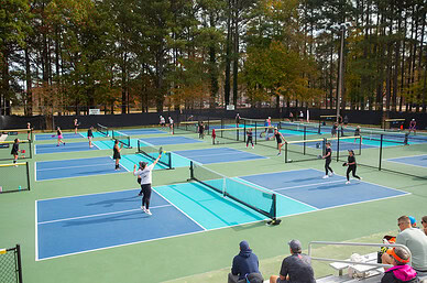Enthusiastic adult pickleball players playing on a hard court surface surrounded by trees with a crowd observing at Pickleball Virginia Beach