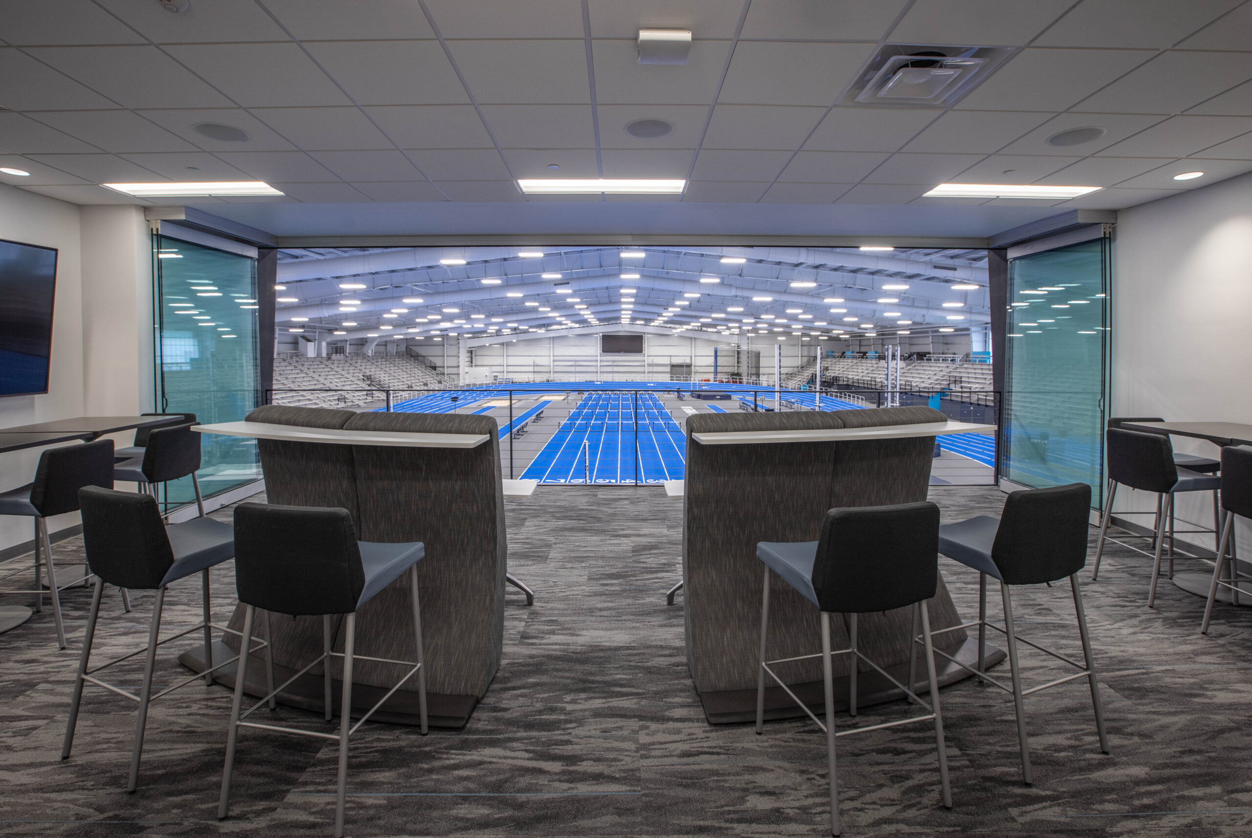 An Indoor view of the Virginia Beach Sports Center track from the second floor suite with a vibrant blue track, metal stands, and field equipment. 