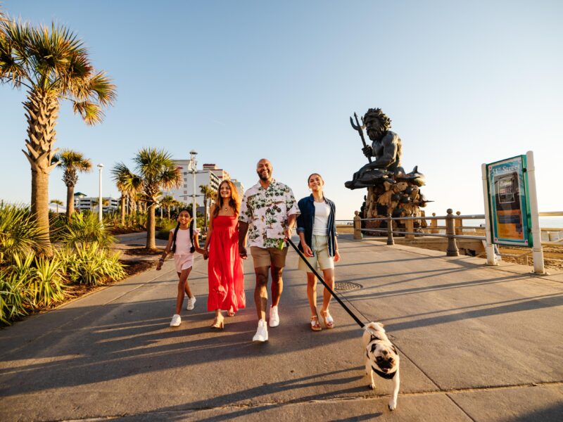 A family with two adults and two children walk along a palm-lined boardwalk, a dog on a leash, with the King Neptune statue in the background.