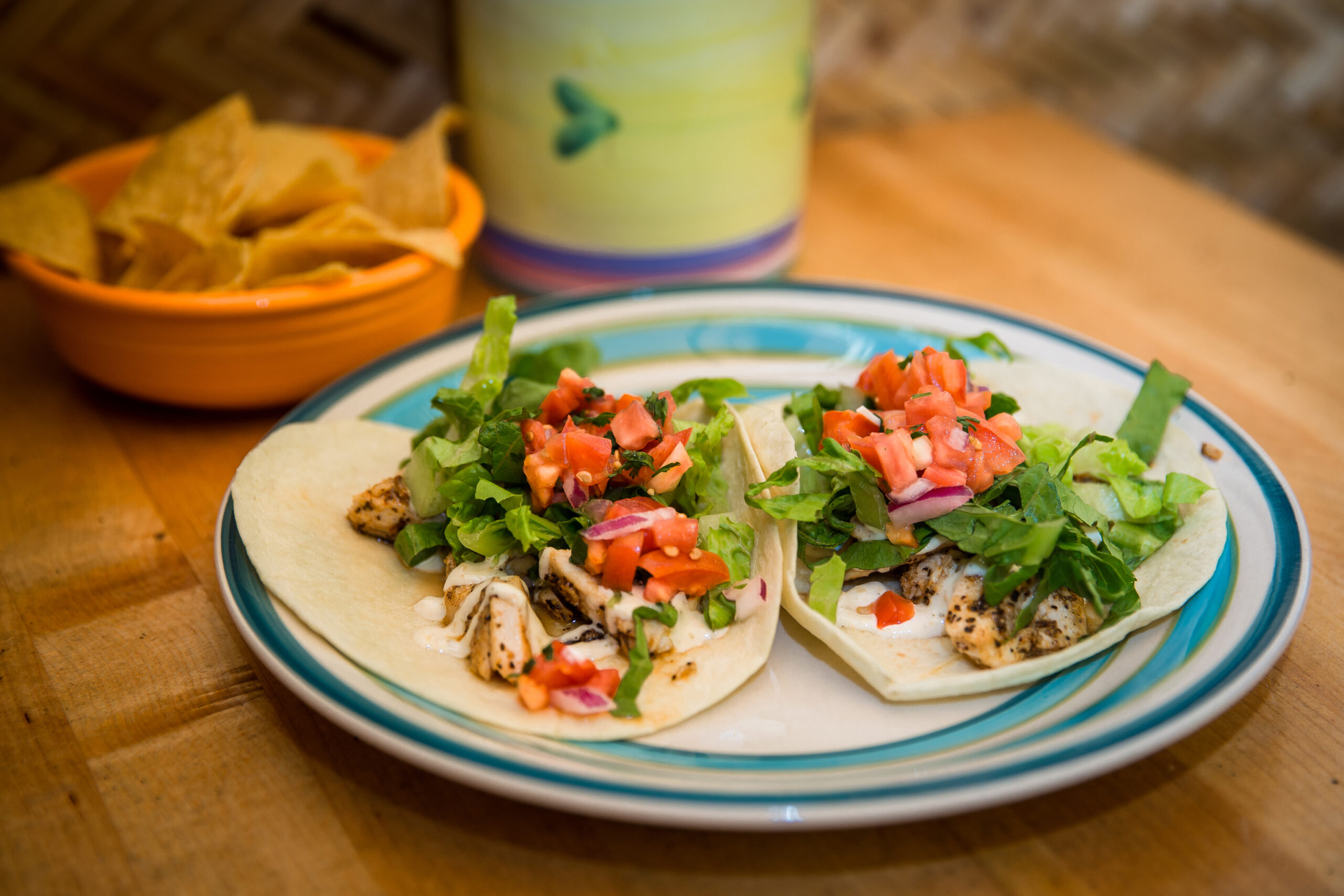 Two soft tacos filled with grilled chicken, lettuce, diced tomatoes, and onions on a plate. A bowl of tortilla chips and a decorative container are in the background.